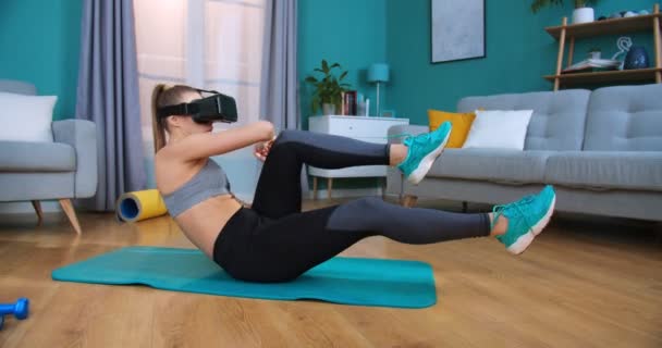 Ajuste mujer joven sentada en la estera de yoga, poniéndose gafas de realidad virtual doingvariation de entrenamiento de fitness en línea. Entrenamiento femenino y ejercicios abdominales flexionar las piernas en el suelo en casa, vista lateral . — Vídeos de Stock