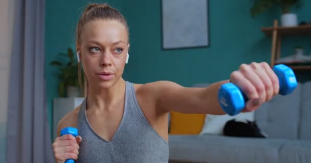 Close up retrato de uma mulher fitness que trabalha com halteres na sala de estar. Mulher concentrada e concentrada a treinar os braços. Desporto e fitness. Conceito de treinamento, treino e bem-estar . — Vídeo de Stock