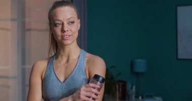 Sevimli, genç ve zinde bir kadın yoğun spor antrenmanından sonra evinde duruyor ve şişeden su içiyor ve kameraya bakıyor. Spor ve spor. Eğitim, egzersiz ve sağlık konsepti.