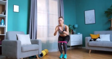 Yetişkin bir kadın direnç bandıyla evde çömeliyor, kalçaları için spor yapıyor. Spor ve spor. Eğitim, egzersiz ve sağlık konsepti.