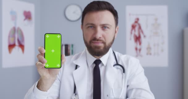 Портрет кавказького лікаря в білій сукні з стетоскопом у його медичному кабінеті, що показує вертикальний смартфон з зеленим екраном. Чоловік-лікар демонструє телефон з хроматичним ключем. Рух стеження — стокове відео