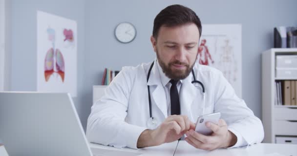 Medium plan van knappe arts surfen op internet tijdens de lunchpauze. Dokter leest nieuws met smartphone. Stagiaire werkt aan de telefoon zittend in het kantoor in witte medische jurk met een statoscoop. — Stockvideo