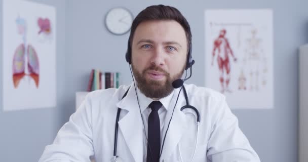 Online-Arztbesuche. Familientherapeut im Kopfhörer sitzt am Tisch im Schrank und berät Patienten per Webcam. Mann im Videochat, ärztliche Beratung während der Quarantäne. — Stockvideo