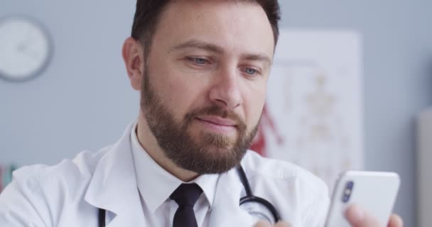 Retrato de cerca del guapo doctor buscando en internet. Asistente de salud leyendo noticias usando smartphone. Terapeuta trabajando en el teléfono sentado en la oficina en bata médica blanca con un statoscopio . — Vídeos de Stock