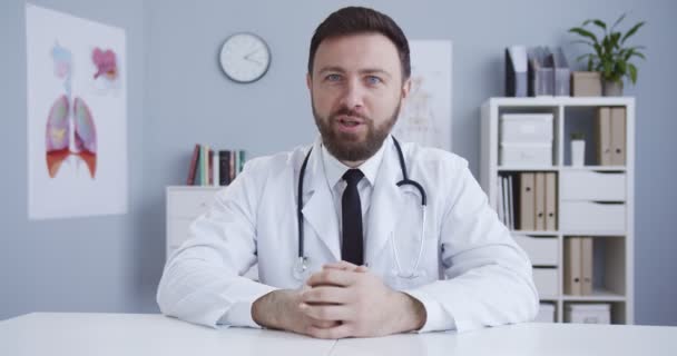 Schöne blauäugige männliche Arzt Blogger in weißer Robe in der Klinik Büro Beratung über Gesundheitsversorgung online. Mann, Krankenhausangestellter mit professioneller medizinischer Beratung im Internet während der Qurantine. — Stockvideo