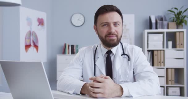 Běloch v nemocničním pokoji, portrét pohledného modrookého muže v bílých šatech se stetoskopem v ordinaci. Zdravotník sedí za stolem. Perfektní lékařská služba na klinice. — Stock video