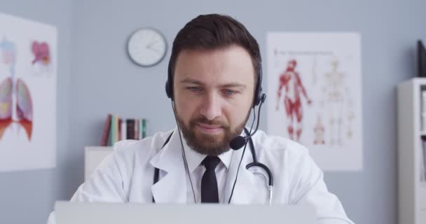网上医生咨询。家庭治疗师的肖像在耳机咨询病人通过网络摄像头使用笔记本电脑。男子与病人进行视频交谈，在检疫期间提供医疗咨询. — 图库视频影像
