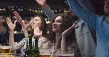 Beyaz erkek ve kadın arkadaşlar, spor maçı izlerken geceyi barda geçirirler. Erkekler ve kadınlar alkışlıyor ve el hareketi yapıyorlar. Futbol taraftarları televizyonda bira içip tezahürat yapıyorlar..