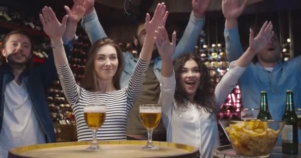Caucásico amigos masculinos y femeninos animando en el bar al ver el juego deportivo. Hombres y mujeres aplaudiendo y haciendo gestos de manos en el pub. Los aficionados al fútbol ven el partido en la televisión con cerveza y aperitivos . — Vídeos de Stock