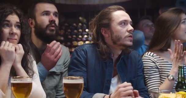 Kaukascy faceci i dziewczęta siedzą wieczorem w pubie z piwem i oglądają mecz sportowy w telewizji. Przyjaciele martwią się o drużynę futbolową w barze. mężczyzna i kobieta smutni i rozczarowani fani. — Wideo stockowe