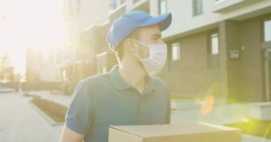 Mavi şapkalı ve tıbbi maskeli bir teslimatçı sokakta yürüyor ve elinde karton kutu taşıyor. Karantina sırasında karton paket teslimatı. Posta konsepti. Evde kal, internetten sipariş ver.