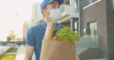 Mavi kep ve tıbbi maske takan bir teslimatçı sokakta yürürken yanında taze yiyecek taşıyan bir paket taşıyor. Karantina sırasında marketten eve kapı kapı teslimat yapan bir kurye. Ürünleri çevrimiçi sipariş