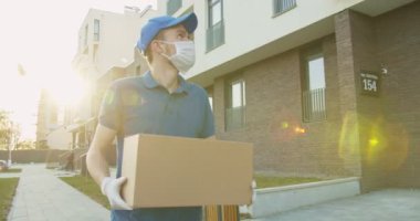 Mavi şapkalı ve tıbbi maskeli bir adam sokakta yürüyor ve elinde karton kutu taşıyor. Karantina sırasında karton paket teslimatı. Posta konsepti. Evde kal, internetten sipariş ver.