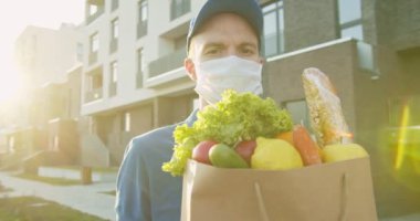 Taze yiyecek paketi taşıyan, kep takan ve tıbbi maske takan teslimatçı portresi. Karantina sırasında kurye eve market, kapı kapı teslimat getiriyor. İnternetten ürün sipariş et, evde kal..