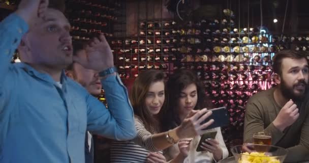 Les gars fans de football regarder match avec des copines le soir dans un pub et réconforter pour l'équipe préférée. Jeunes hommes caucasiens célébrant le résultat du jeu dans le bar. Les filles prennent des photos selfie sur les téléphones . — Video