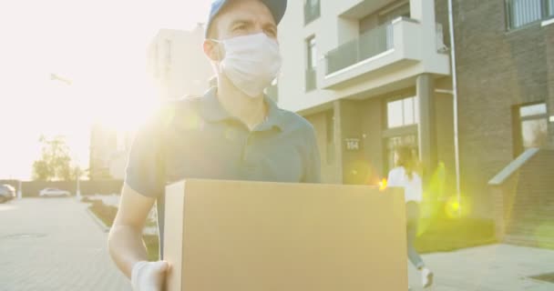 Trabalhador de entregas usando boné e máscara médica andando na rua e carregando caixa de papelão nas mãos. Correio masculino entregando pacote de caixa de papelão durante a quarentena. Concepção de correio. Fique em casa, encomendar online — Vídeo de Stock