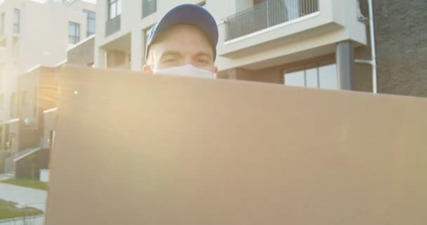 Porträt eines jungen kaukasischen Mannes in blauer Uniform, Hut und medizinischer Maske, der mit einem Paket in der Hand auf der Straße steht. Kurierdienst versendet Kartonpakete während der Quarantäne. Zuhause bleiben, online bestellen. — Stockvideo