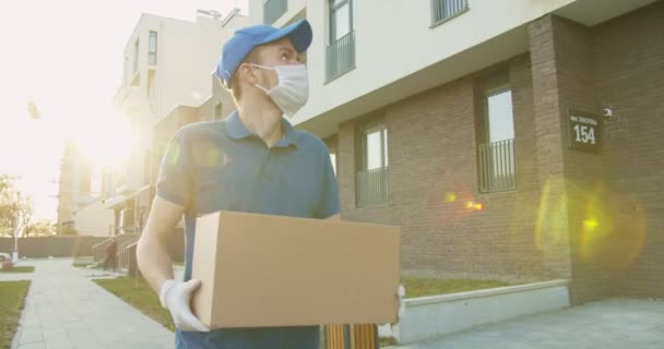 Hombre trabajador de entrega con gorra azul y máscara médica caminando por la calle y llevando caja de cartón en las manos. Mensajero entregando paquete de cartón durante la cuarentena. Concepto de correo. Quedarse en casa, ordenar en línea — Vídeos de Stock