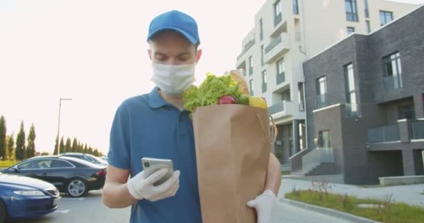 Kurir bertopi biru memegang paket makanan dan mengetuk, mengirim pesan di smartphone outdoor dan mencari alamat. Pria membawa belanjaan ke rumah, pintu ke pintu selama karantina. — Stok Video