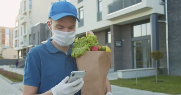 Mensajero de hombre en sombrero azul sosteniendo paquete de alimentos y tocando, mensaje de texto en el teléfono inteligente al aire libre y en busca de dirección. Hombre trayendo comestibles a casa, entrega puerta a puerta durante la cuarentena . — Vídeos de Stock