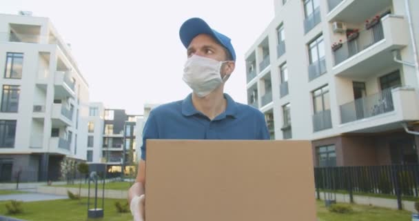 Repartidor con gorra azul y máscara médica caminando por la calle y llevando caja de cartón en las manos. Mensajero entregando paquete de cartón durante la cuarentena. Concepto de correo. Quedarse en casa, ordenar en línea — Vídeos de Stock