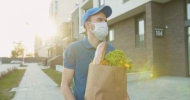 Sağlık maskesi takan bir teslimatçı sokakta yürüyor ve yanında yiyecek paketi taşıyor. Karantina sırasında kurye eve market, kapı kapı teslimat getiriyor. Evde kal, internetten ürün sipariş et..
