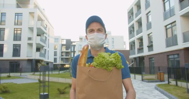 Mavi kep ve tıbbi maske takan bir teslimatçı sokakta yürürken yanında taze yiyecek taşıyan bir paket taşıyor. Karantina sırasında marketten eve kapı kapı teslimat yapan bir kurye. Ürünleri çevrimiçi sipariş — Stok video