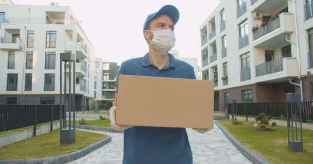 Hombre trabajador de entrega con gorra azul y máscara médica caminando por la calle y llevando caja de cartón en las manos. Mensajero entregando paquete de cartón durante la cuarentena. Concepto de correo. Quedarse en casa, ordenar en línea — Vídeos de Stock