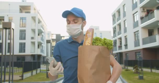 Mensajero de hombre en sombrero azul sosteniendo paquete de alimentos y tocando, mensaje de texto en el teléfono inteligente al aire libre y en busca de dirección. Hombre trayendo comestibles a casa, entrega puerta a puerta durante la cuarentena . — Vídeos de Stock