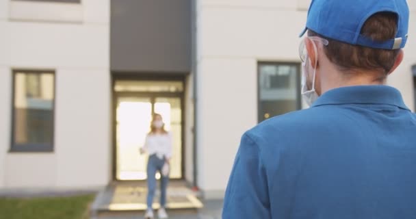 Rear of delivery man walking from supermarket coming to building and meeting young Caucasian woman. Girl in medical mask and gloves handling packet with food from man. Pandemic concept. — Stock Video