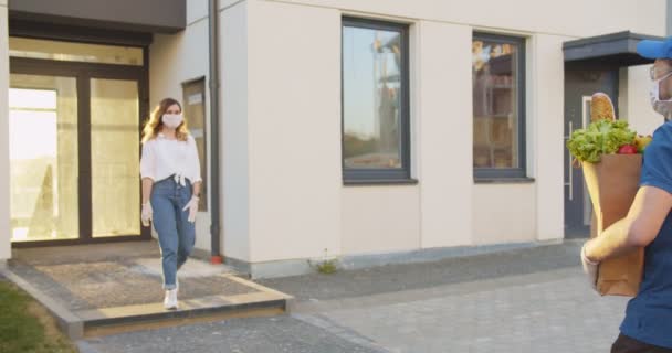 Zijaanzicht van de bezorger die van supermarkt naar gebouw loopt en een jonge blanke vrouw ontmoet. Meisje met medisch masker en handschoenen die pakje hanteren met voedsel van koerier man. Pandemisch concept. — Stockvideo