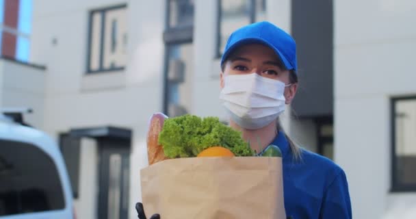 Portrait of delivery woman wearing cap and medical mask carrying packet with fresh food. Courier bringing grocery to house, door to door delivery during quarantine. Order products online stay at home — Stock Video