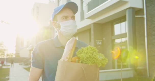 Man delivery worker wearing blue cap and medical mask walking the street and carrying packet with fresh food. Courier bringing grocery to house during quarantine. Stay home, order products online. — Stock Video