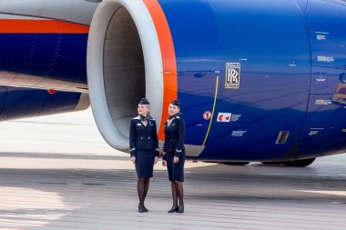 Russia, Vladivostok, 10/13/2017. Beautiful stewardesses dressed in official dark blue uniform of Aeroflot Airlines stand near at Rolls Royce's engine of jet aircraft. Crew of plane. Aviation and transportation. clipart