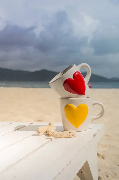 Sabah Beyaz ahşap masa ve plaj arka plan ve güneşli ışık yanında mercan kırmızı ve sarı kalp ile iki su bardağı koymak