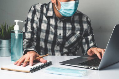 Coronavirus hastalığı veya COVID-19 koruma kavramı - Genç adam, Corona virüsüne karşı korunmak için masadaki dizüstü bilgisayarı kullanırken koruyucu ve temizlik ekipmanlarıyla evde çalışıyor.
