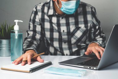 Coronavirus hastalığı veya COVID-19 koruma kavramı - Genç adam, Corona virüsüne karşı korunmak için masadaki dizüstü bilgisayarı kullanırken koruyucu ve temizlik ekipmanlarıyla evde çalışıyor.