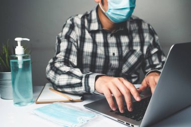 Coronavirus hastalığı veya COVID-19 koruma kavramı - Genç adam, Corona virüsüne karşı korunmak için masadaki dizüstü bilgisayarı kullanırken koruyucu ve temizlik ekipmanlarıyla evde çalışıyor.