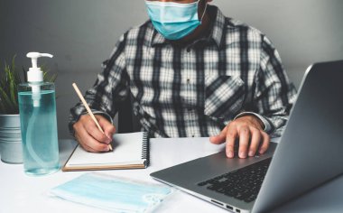 Coronavirus hastalığı veya COVID-19 koruma kavramı - Genç adam, Corona virüsüne karşı korunmak için masadaki dizüstü bilgisayarı kullanırken koruyucu ve temizlik ekipmanlarıyla evde çalışıyor.