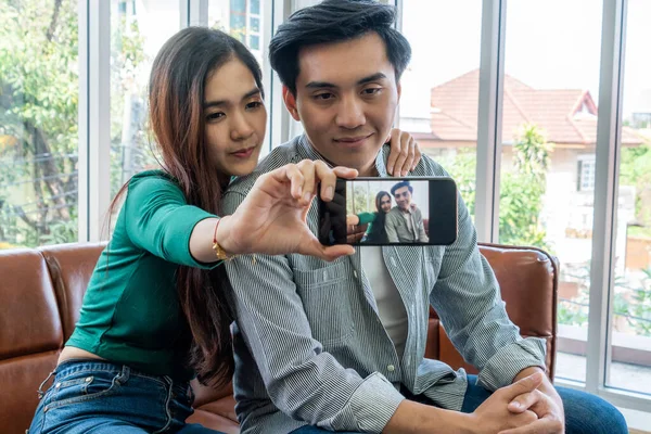 幸せなアジアのカップルは自宅のリビングルームで携帯電話で自撮り写真を撮るのを楽しむ — ストック写真