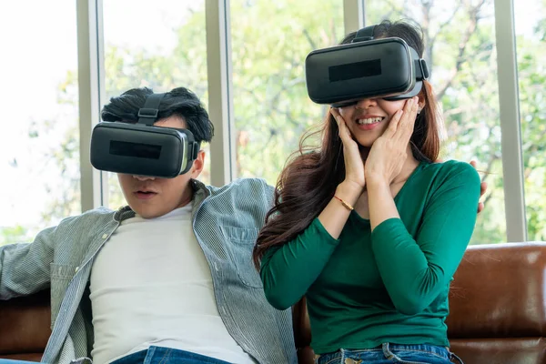 Happy couple watching video in virtual reality VR headset in living room at home. Multimedia movie and entertainment concept.