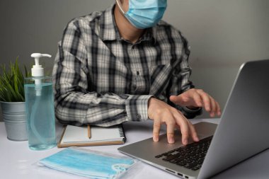 Coronavirus hastalığı veya COVID-19 koruma kavramı - Genç adam, Corona virüsüne karşı korunmak için masadaki dizüstü bilgisayarı kullanırken koruyucu ve temizlik ekipmanlarıyla evde çalışıyor.