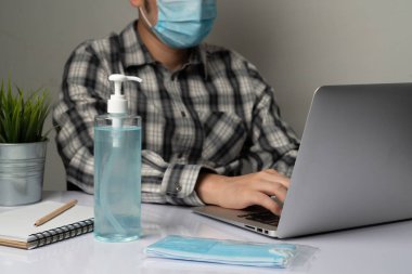 Coronavirus hastalığı veya COVID-19 koruma kavramı - Genç adam, Corona virüsüne karşı korunmak için masadaki dizüstü bilgisayarı kullanırken koruyucu ve temizlik ekipmanlarıyla evde çalışıyor.