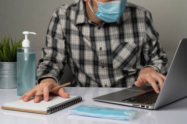 Coronavirus hastalığı veya COVID-19 koruma kavramı - Genç adam, Corona virüsüne karşı korunmak için masadaki dizüstü bilgisayarı kullanırken koruyucu ve temizlik ekipmanlarıyla evde çalışıyor.