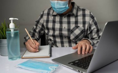 Coronavirus hastalığı veya COVID-19 koruma kavramı - Genç adam, Corona virüsüne karşı korunmak için masadaki dizüstü bilgisayarı kullanırken koruyucu ve temizlik ekipmanlarıyla evde çalışıyor.