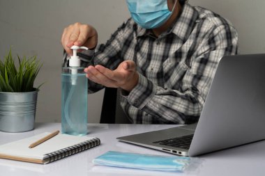 Coronavirus hastalığı veya COVID-19 koruma kavramı - Genç adam, Corona virüsüne karşı korunmak için masadaki dizüstü bilgisayarı kullanırken koruyucu ve temizlik ekipmanlarıyla evde çalışıyor.