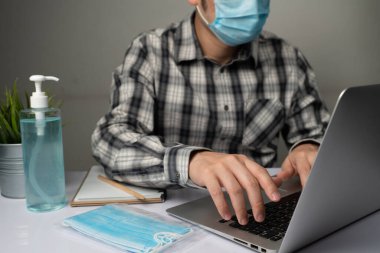 Coronavirus hastalığı veya COVID-19 koruma kavramı - Genç adam, Corona virüsüne karşı korunmak için masadaki dizüstü bilgisayarı kullanırken koruyucu ve temizlik ekipmanlarıyla evde çalışıyor.