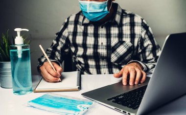 Coronavirus hastalığı veya COVID-19 koruma kavramı - Genç adam, Corona virüsüne karşı korunmak için masadaki dizüstü bilgisayarı kullanırken koruyucu ve temizlik ekipmanlarıyla evde çalışıyor.