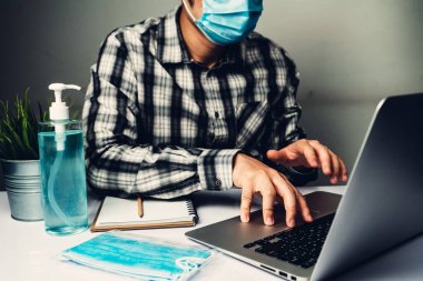 Coronavirus hastalığı veya COVID-19 koruma kavramı - Genç adam, Corona virüsüne karşı korunmak için masadaki dizüstü bilgisayarı kullanırken koruyucu ve temizlik ekipmanlarıyla evde çalışıyor.