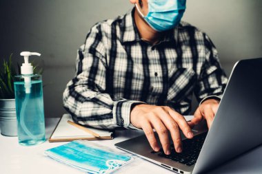 Coronavirus hastalığı veya COVID-19 koruma kavramı - Genç adam, Corona virüsüne karşı korunmak için masadaki dizüstü bilgisayarı kullanırken koruyucu ve temizlik ekipmanlarıyla evde çalışıyor.